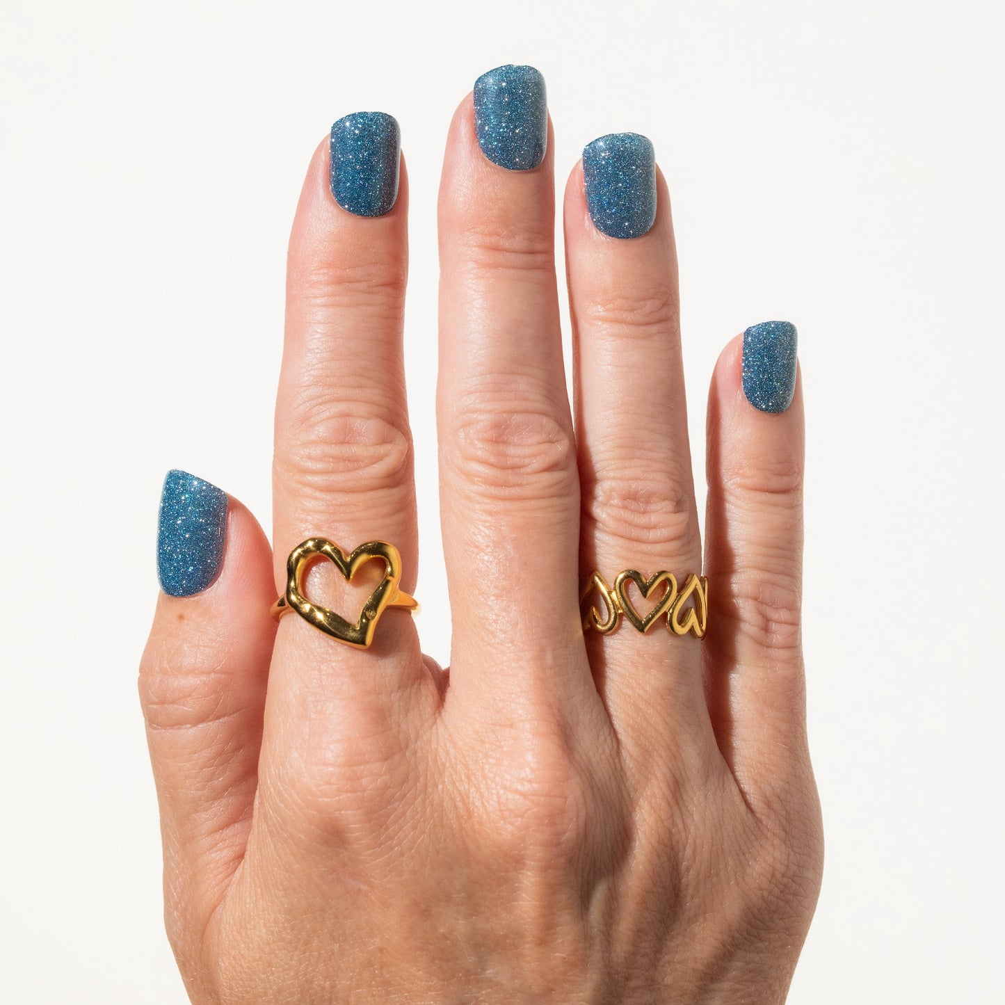 Hand wearing two gold heart-shaped rings with blue glitter press on nails on a white background