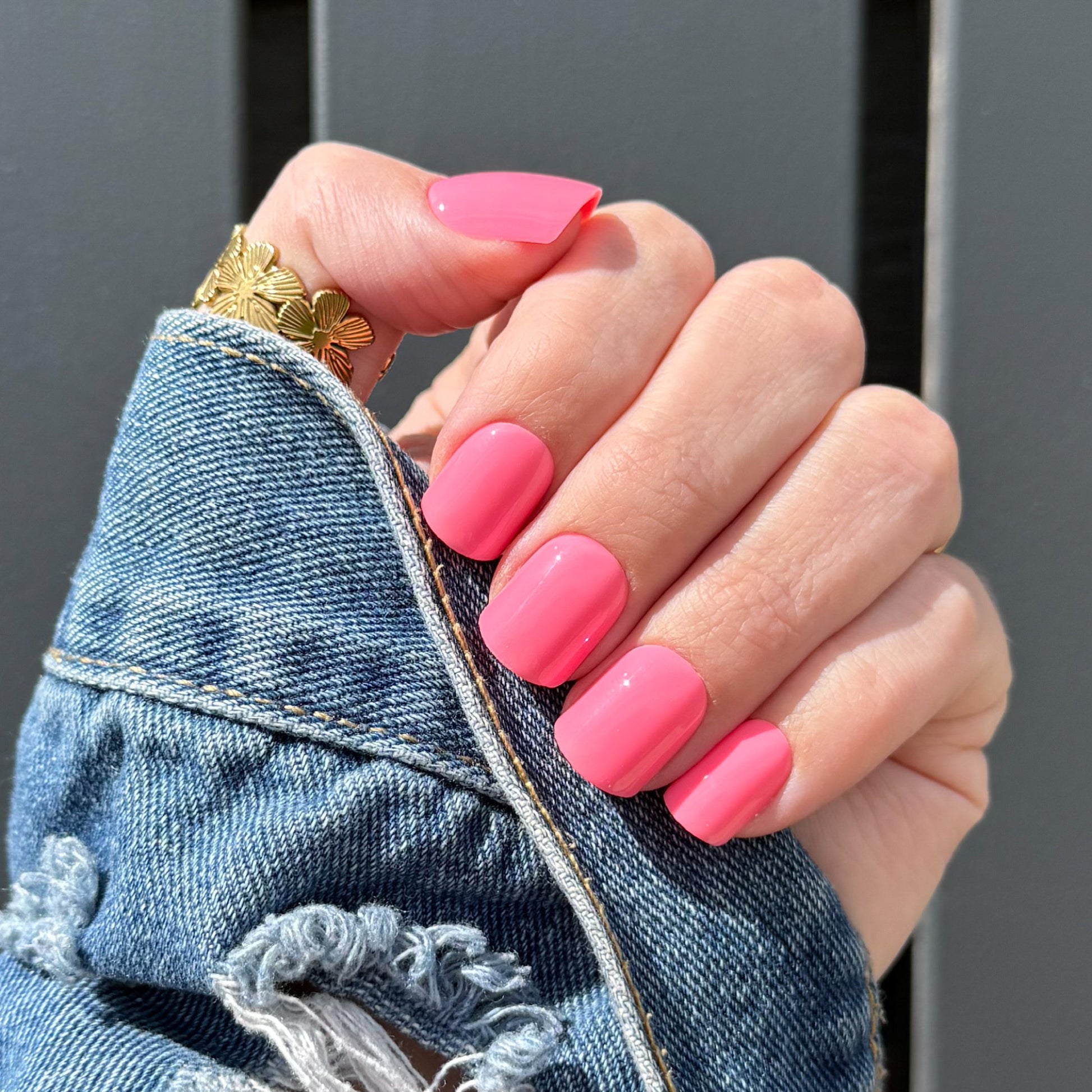 Hand with pink nails wearing a gold ring, against a gray background