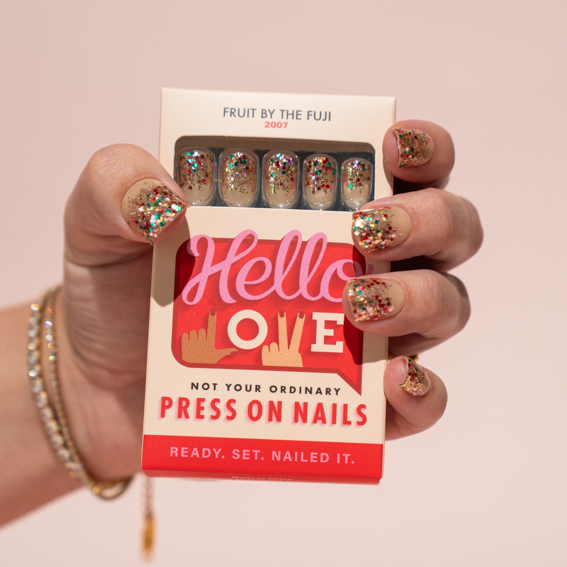 Box of Hello Love press-on nails held by a hand with glittery press on nails against a pink background