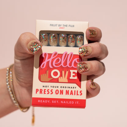 Box of Hello Love press-on nails held by a hand with glittery press on nails against a pink background