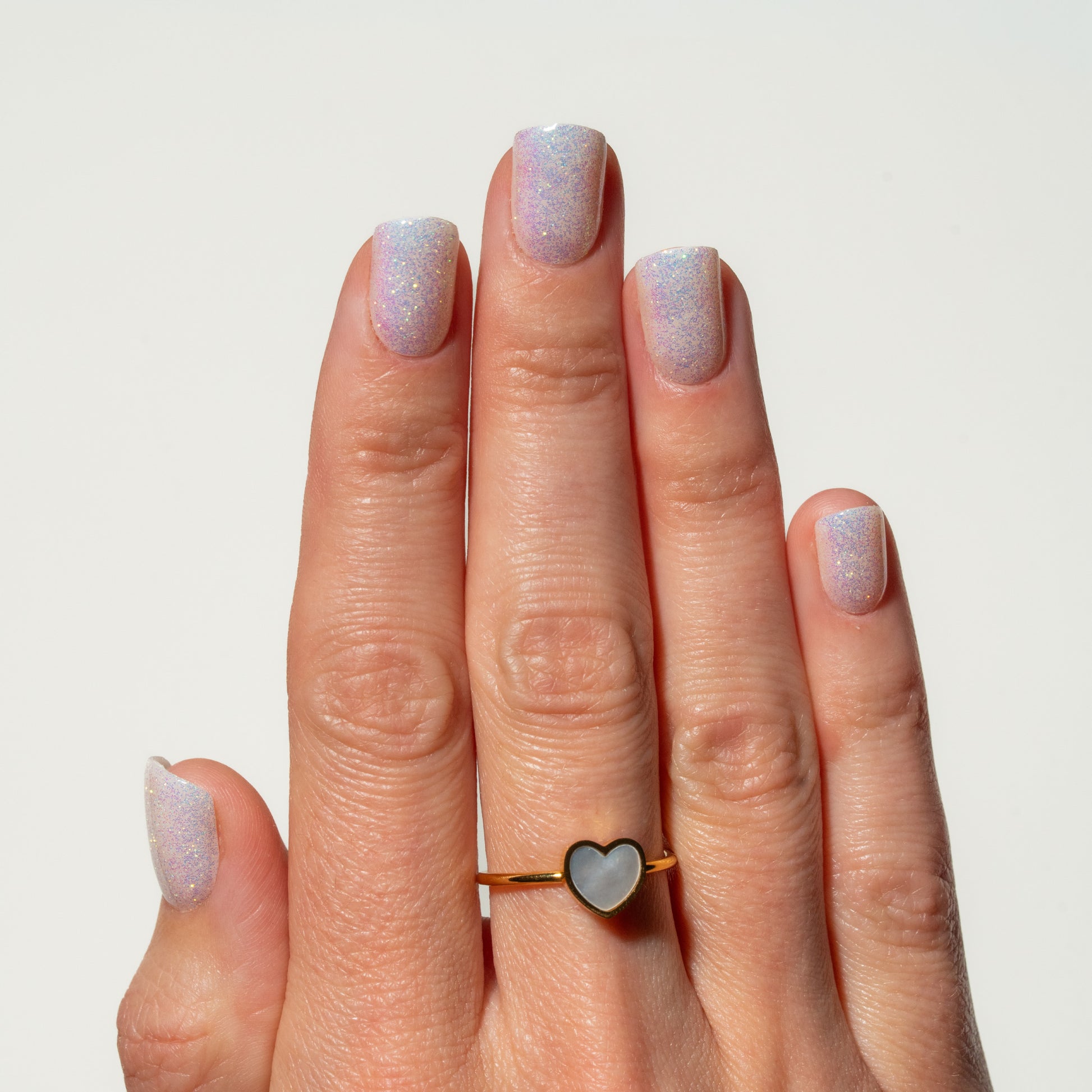 Hand with glittery nails wearing a heart-shaped ring on a plain background