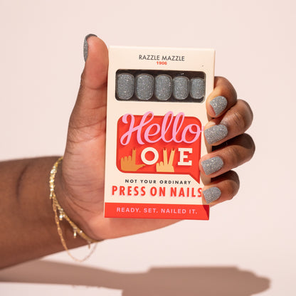 Hand holding a box of Hello Love press-on nails with silver glitter nails.