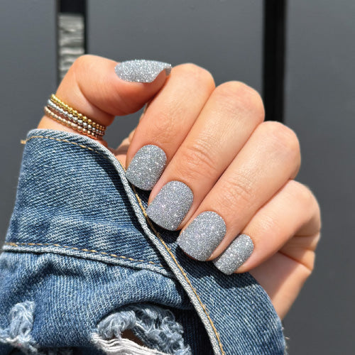 Hand with glittery silver nails wearing a denim jacket against a gray background
