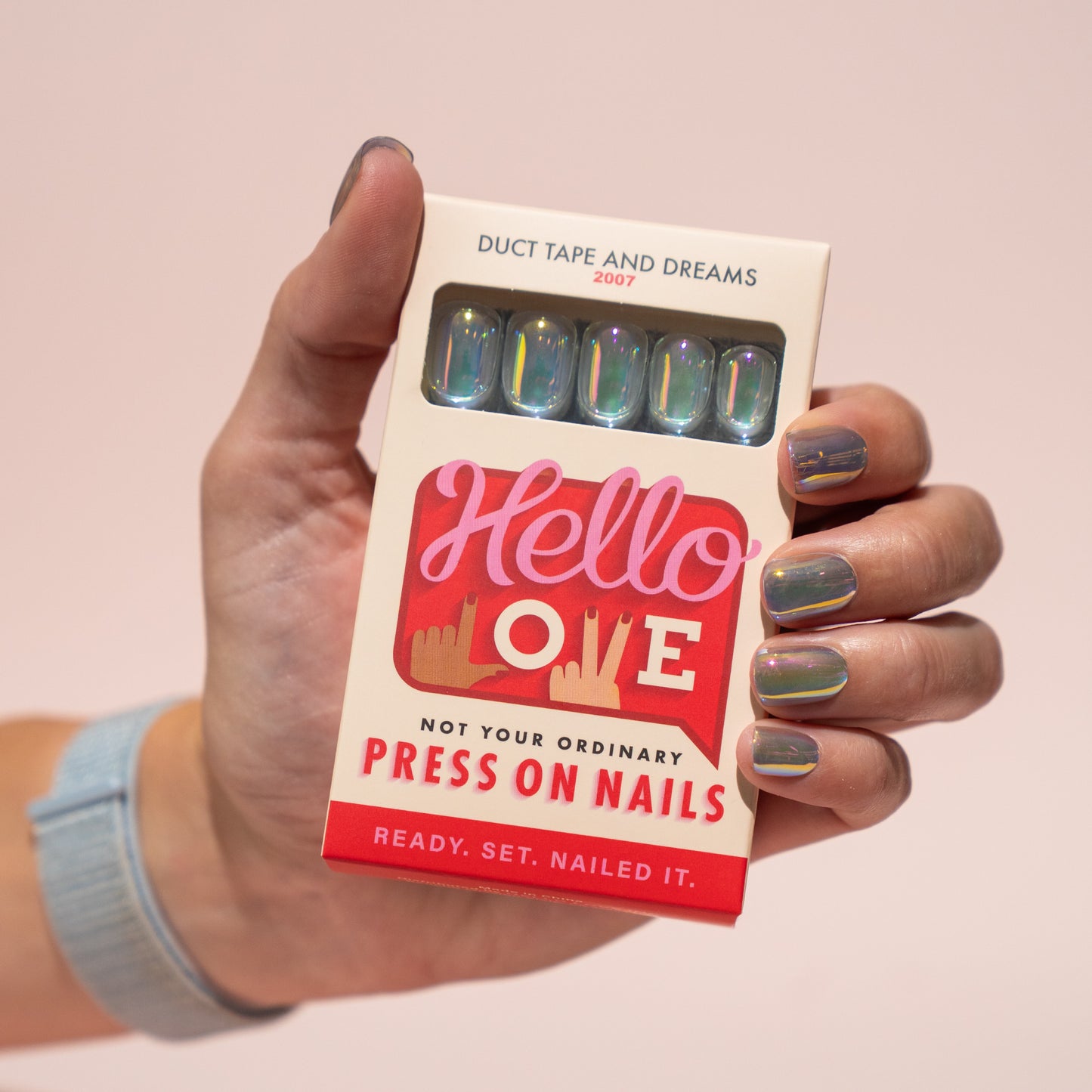 Hand holding a box of 'Hello Love' press-on nails with a pink background