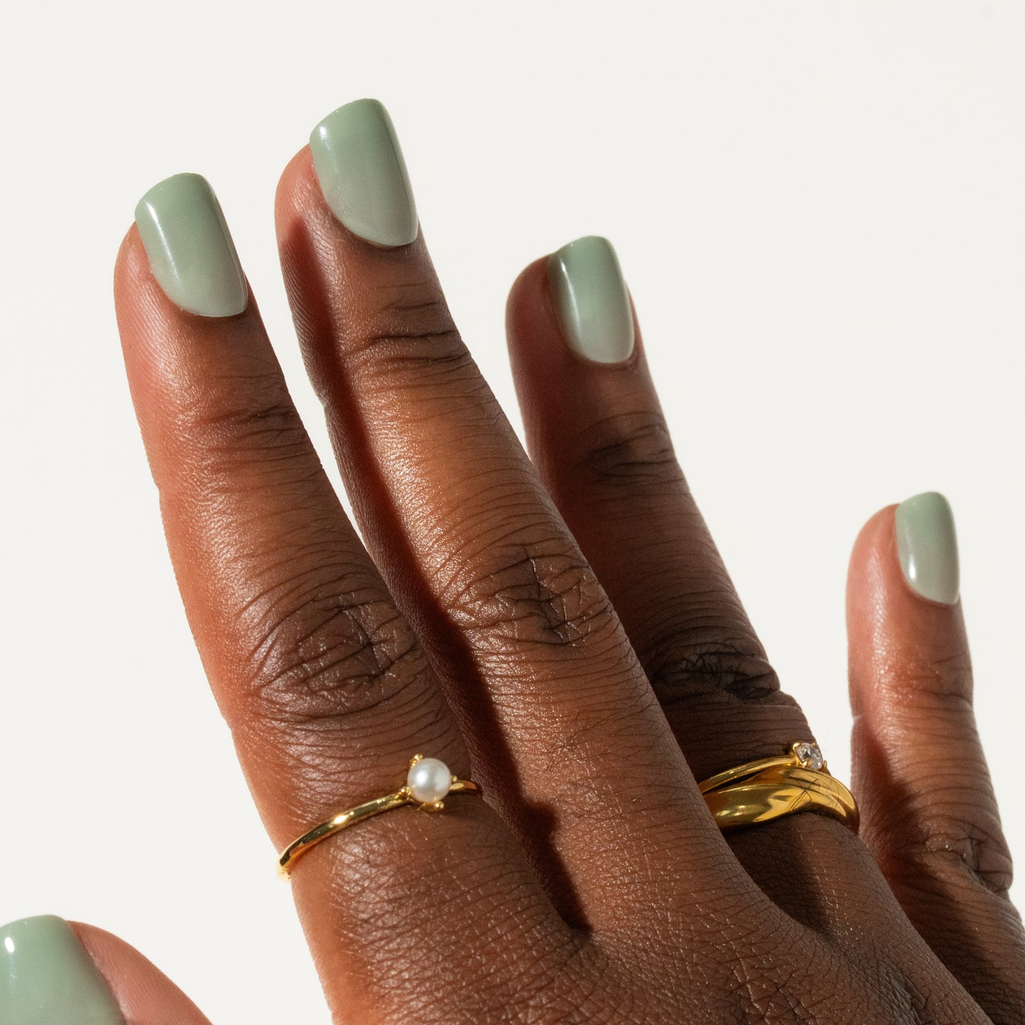 Close-up of a hand with green press on nails wearing two gold rings on a neutral background