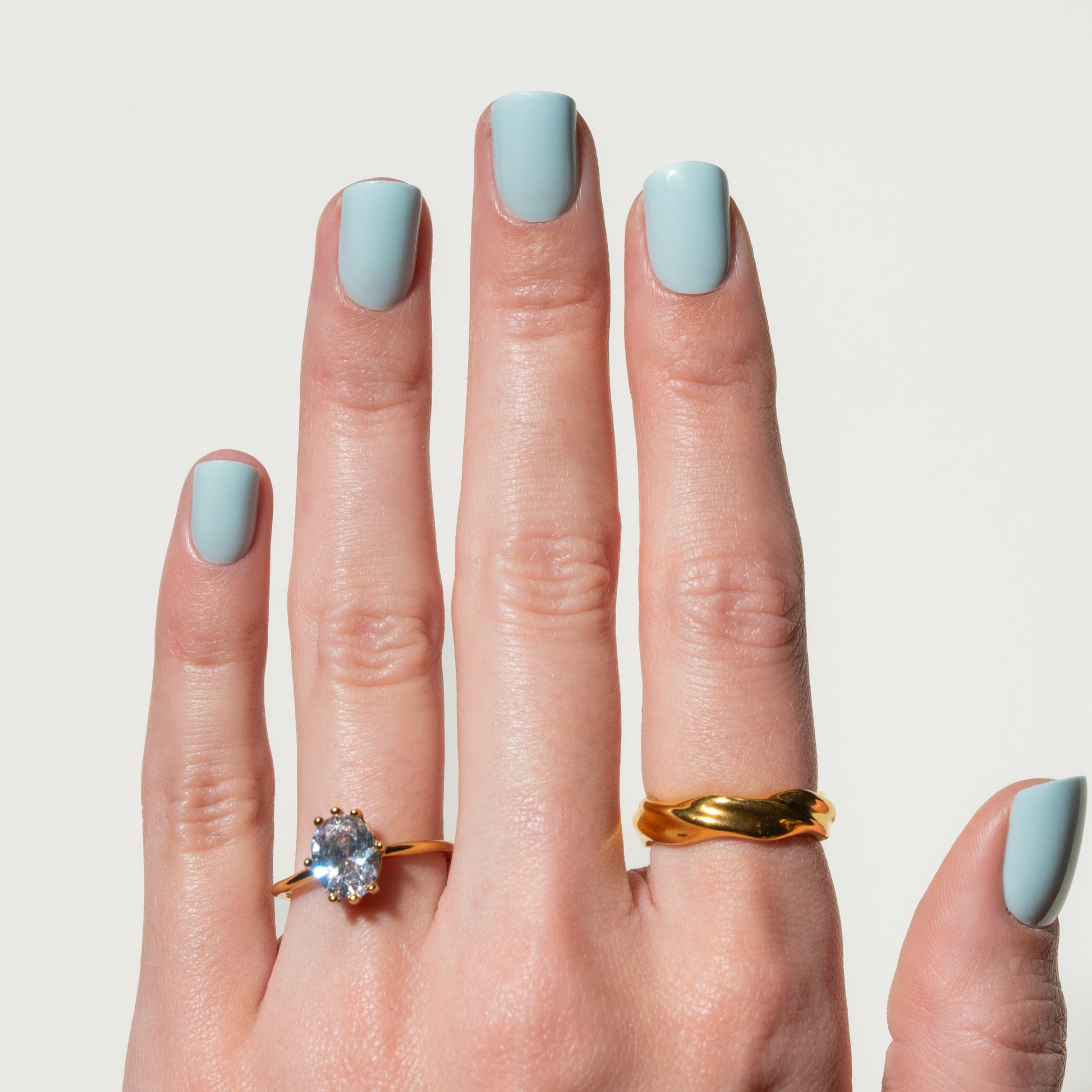 Hand with light blue press on nails wearing two gold rings on a plain background