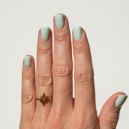 Hand with mint green nails wearing a gold ring on a light gray background