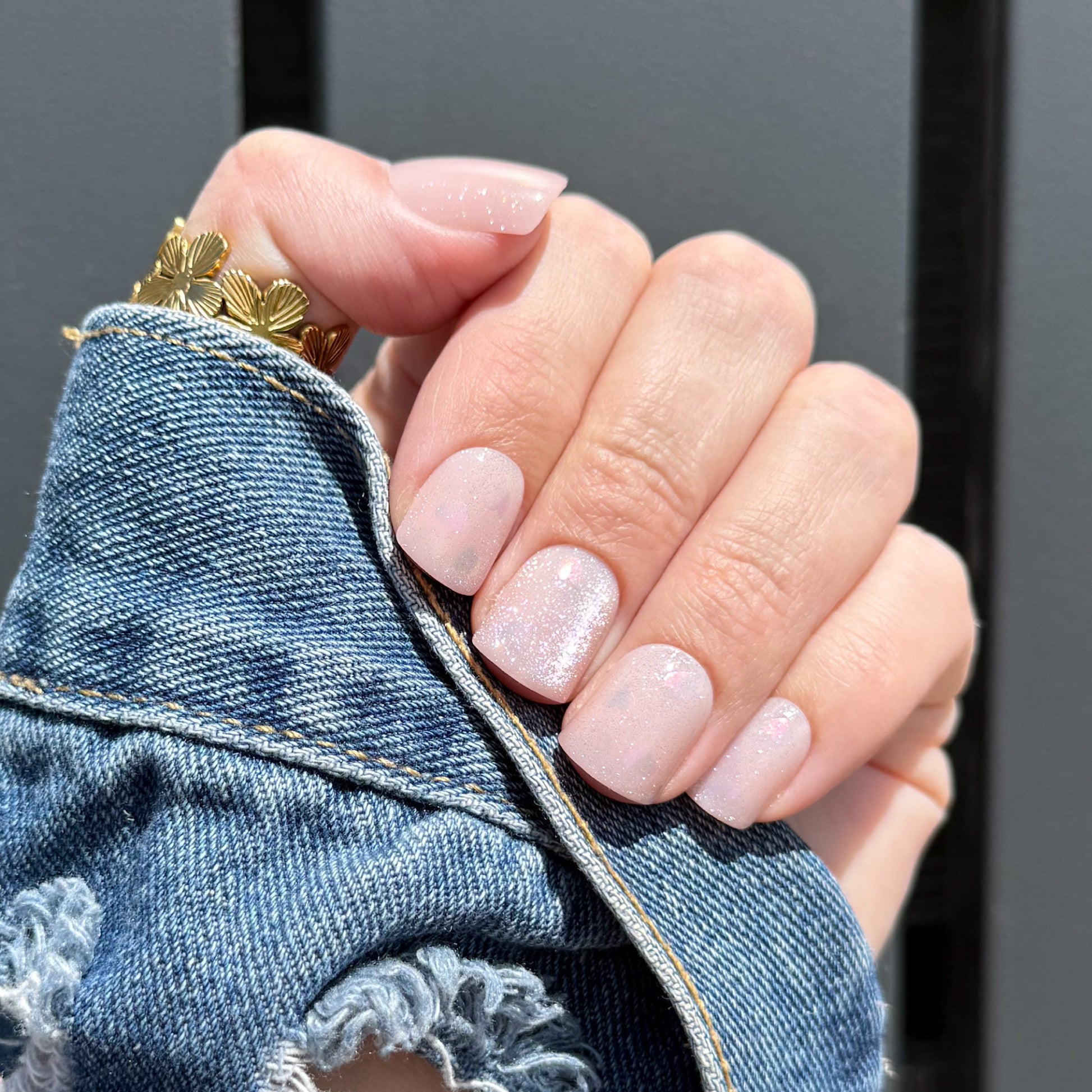 Close-up of a hand with pink press on nails wearing a gold ring, holding denim fabric.