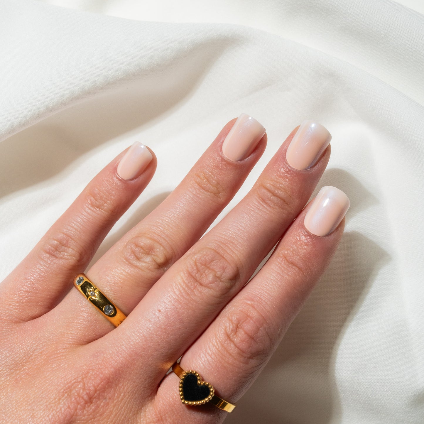 Hand with beige press on nails wearing two gold rings on a white fabric background