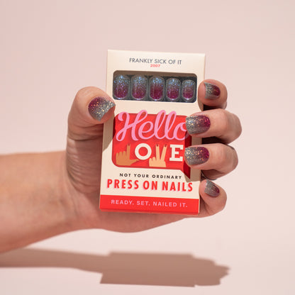 Hand holding a box of Hello Love press-on nails with glittery nails against a plain background