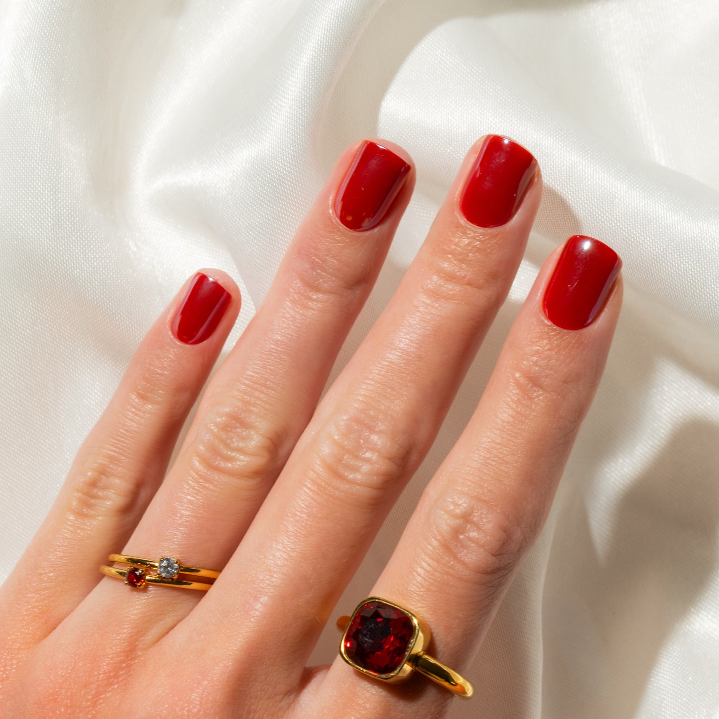 Hand with red press on nails wearing two gold rings on a white fabric background