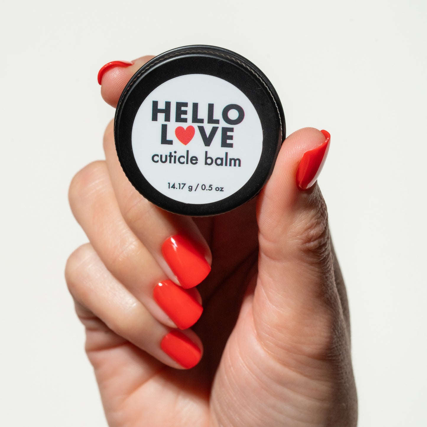 Hand holding a small container of 'Hello Love' cuticle balm with red press on nails on a light background