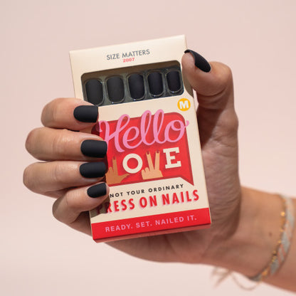 Hand holding a box of black press on nails with 'Hello Love' branding on a light pink background