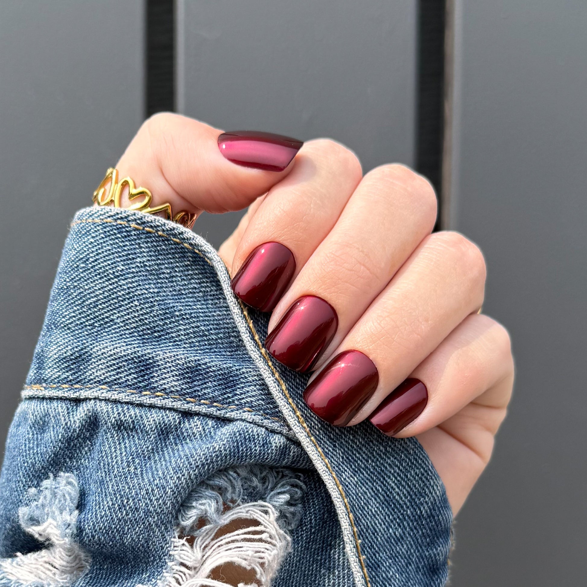 A close-up of a hand with glossy metallic deep burgundy nails, wearing a gold ring and holding the cuff of a distressed blue denim jacket against a dark gray background.