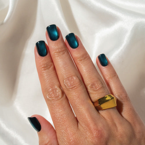 Hand with teal press on nails and a gold ring on a white fabric background