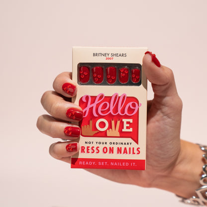 Hand holding a box of 'Hello Love' press on nails with red nails on a beige background